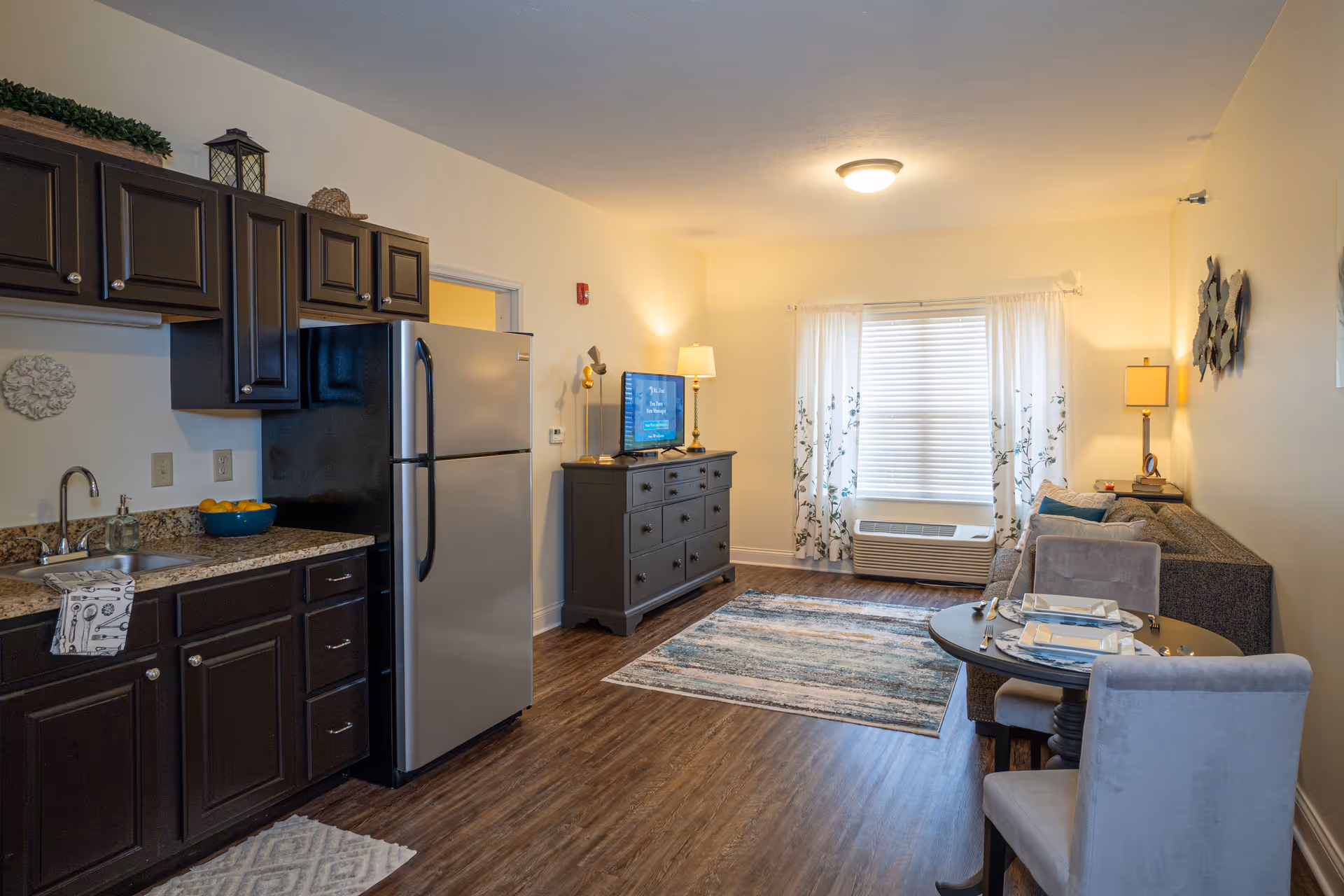 A cozy living space featuring a small kitchen area with dark wood cabinets, a stainless steel refrigerator, and a sink. Adjacent to the kitchen is a living room with a gray sofa, a TV on a dark dresser, a window with white curtains, and a small round dining table set for two with gray upholstered chairs. The room has wooden flooring and soft lighting from lamps.