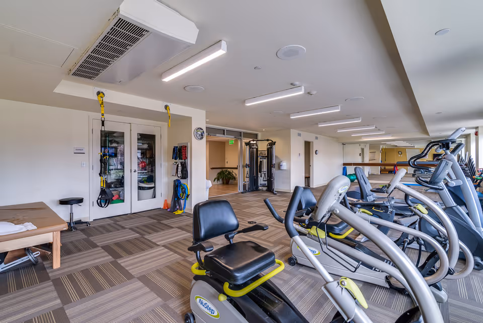 A spacious fitness room with exercise equipment including stationary bikes, elliptical machines, and resistance training equipment. The room has carpeted flooring, bright overhead lighting, and large windows letting in natural light. There are also exercise bands and other fitness accessories hanging on the wall.