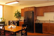 Communal dining area with a wooden table and chairs beside a kitchenette featuring wooden cabinets and black appliances.