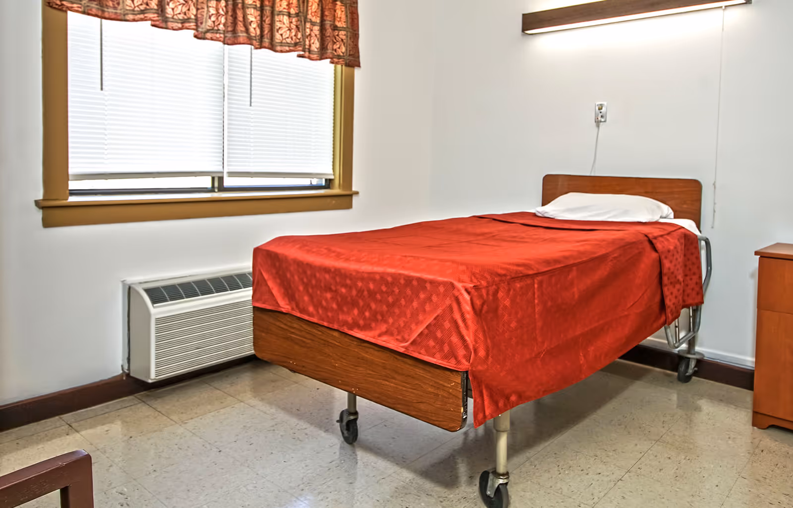 A simple room with a single hospital-style bed covered with a red blanket and a white pillow. The bed has wheels and a wooden headboard. There is a window with closed blinds and a patterned valance above it. Below the window is a wall-mounted air conditioning unit. A wooden nightstand is visible to the right of the bed. The floor is tiled and the walls are plain white.