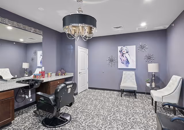 A stylish salon room with a black salon chair in front of a large mirror and granite countertop. The walls are painted purple and decorated with modern art and wall sculptures. There are two white tufted chairs, a small side table with a lamp and plant, and a patterned carpeted floor. A black and silver chandelier hangs from the ceiling.