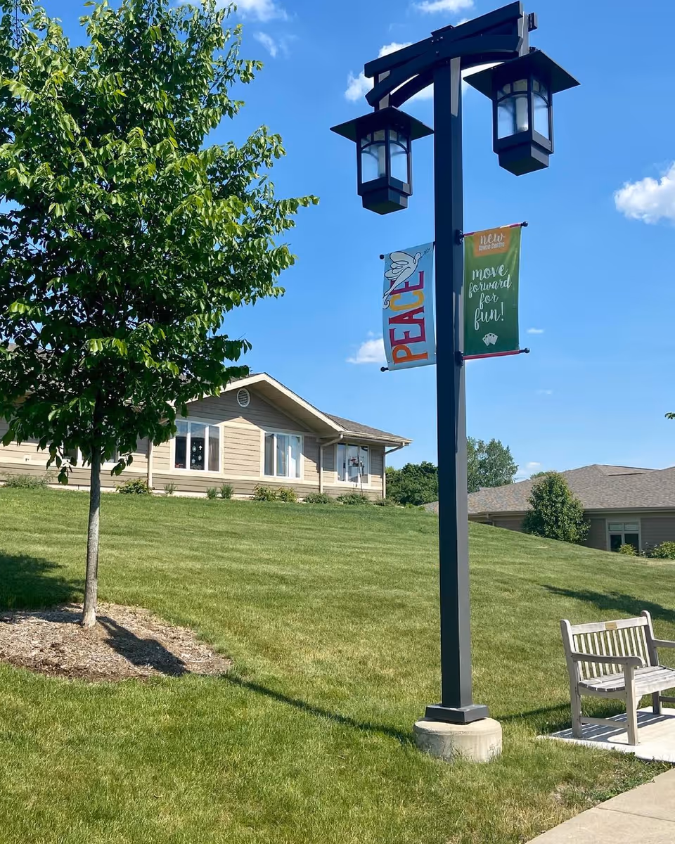 Outdoor scene at The Marquardt facility showing a grassy lawn with a tree, a black lamp post with two lanterns and colorful banners that read 'PEACE' and 'move forward for fun!', a wooden bench on a concrete pad, and beige single-story buildings in the background under a clear blue sky.