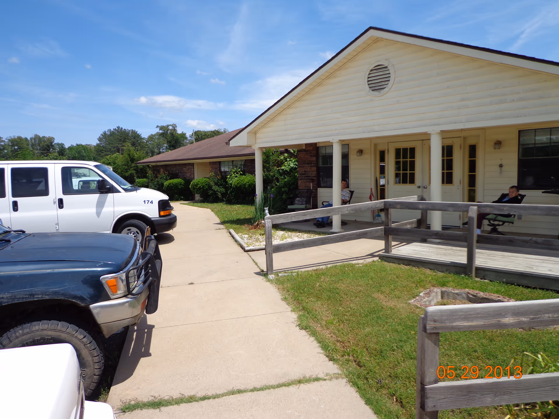 Exterior view of a single-story building with a covered porch and ramp. Two people are sitting on chairs on the porch. There are two vehicles parked on the driveway in front of the building. The sky is clear with a few clouds.