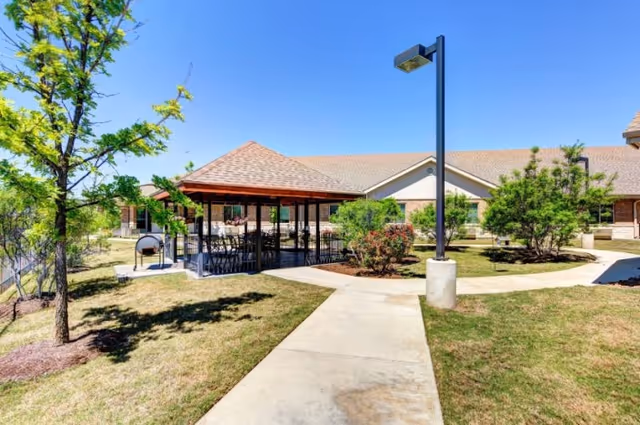 Outdoor area of Hunters Pond Rehabilitation and Healthcare Center featuring a paved walkway leading to a covered pavilion with tables and chairs, surrounded by grass, trees, shrubs, and a building in the background under a clear blue sky.