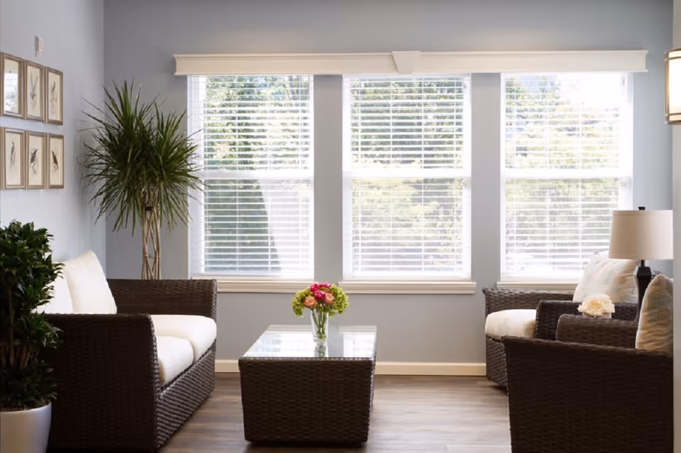 Sunlit sitting area with wicker sofas and chairs around a coffee table topped with flowers in front of three large windows.