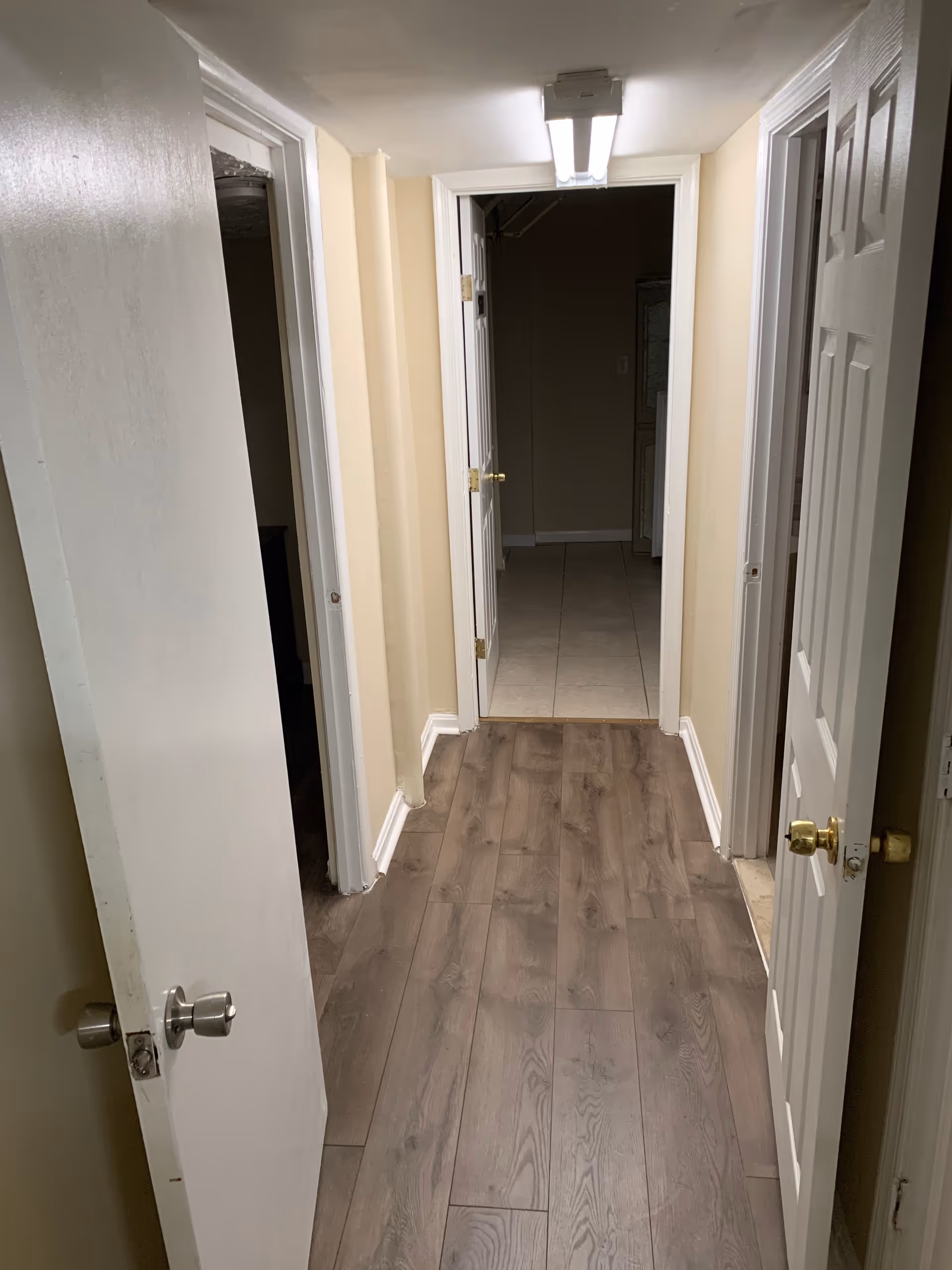 A narrow hallway with light beige walls and wood-patterned flooring. There are three white doors partially open on either side of the hallway, and a fluorescent ceiling light fixture is visible at the end of the hallway.