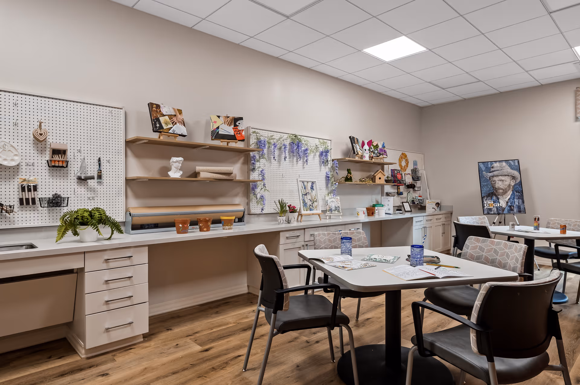 A well-lit craft room with tables and chairs arranged for activities. The walls have pegboards and shelves displaying various craft supplies, decorations, and artwork. The floor is wooden, and the ceiling has white tiles. There are coloring sheets and cups on the tables.