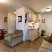 Interior view of a senior living facility showing a cozy seating area with two armchairs and a sofa. The room features wood flooring, light-colored walls, and a small kitchenette area with cabinets and a countertop. There are framed pictures on the walls and a door leading to another room or hallway.