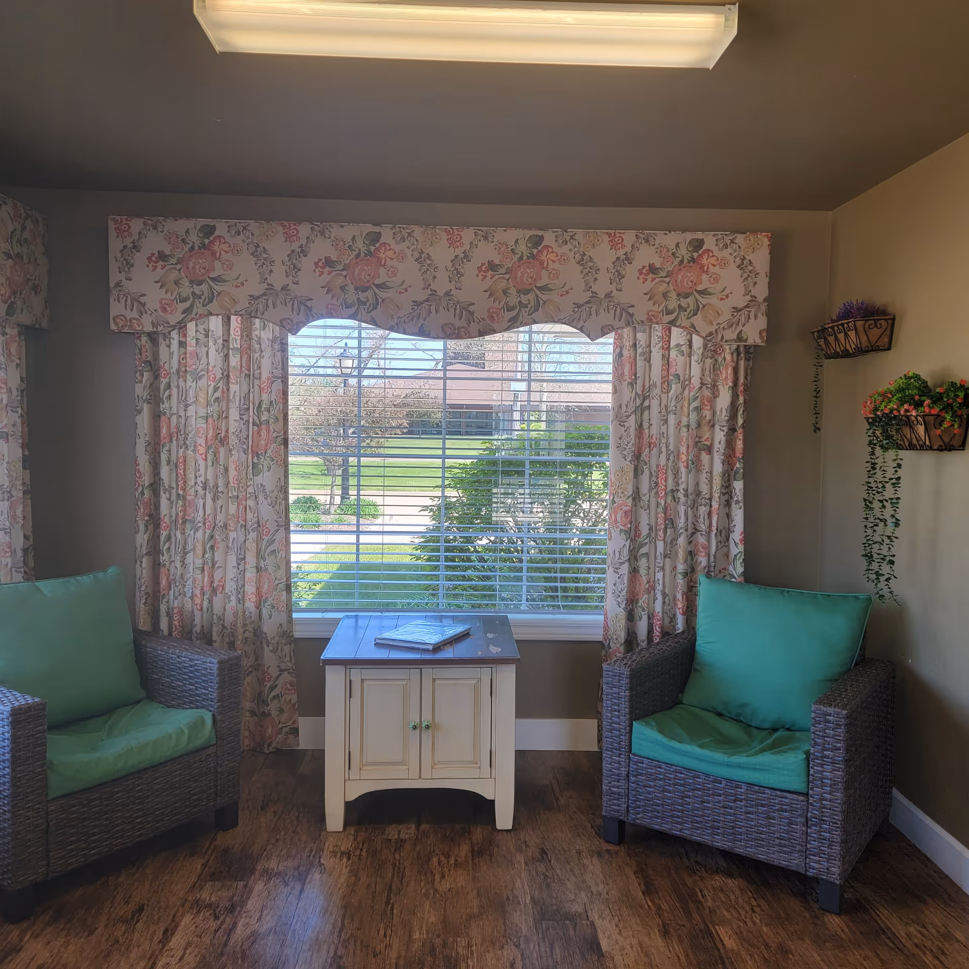 A cozy sitting area with two wicker armchairs featuring green cushions, positioned on either side of a small white wooden cabinet. Behind the cabinet is a large window with floral-patterned curtains and valance, showing a view of a green lawn and trees outside. The walls are beige, and there are small wall-mounted baskets with artificial plants on the right side.