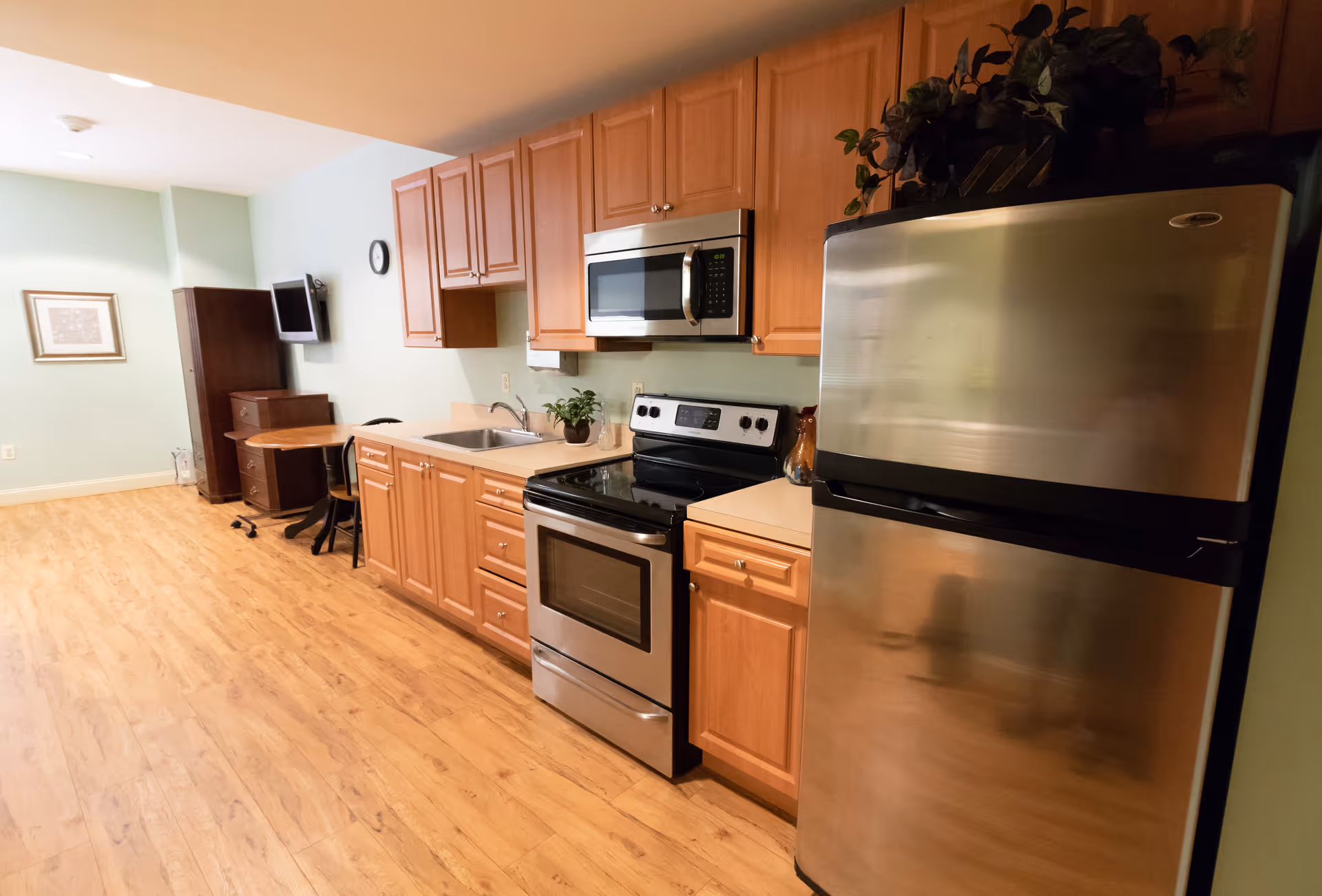 A kitchen area with wooden cabinets, a stainless steel refrigerator, stove, and microwave. There is a sink with a small plant on the counter. The floor is wooden, and in the background, there is a small table with chairs, a wall-mounted TV, and a framed picture on a light green wall.