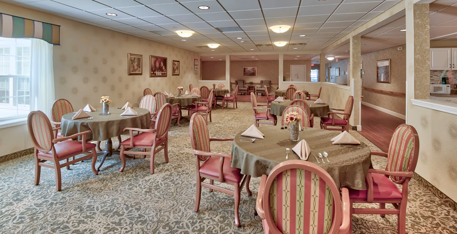 A dining room in an assisted living facility with round tables covered in brown tablecloths, each set with folded napkins, silverware, and small flower arrangements. The room has patterned carpet, striped upholstered chairs, framed artwork on the walls, and large windows with sheer curtains allowing natural light.