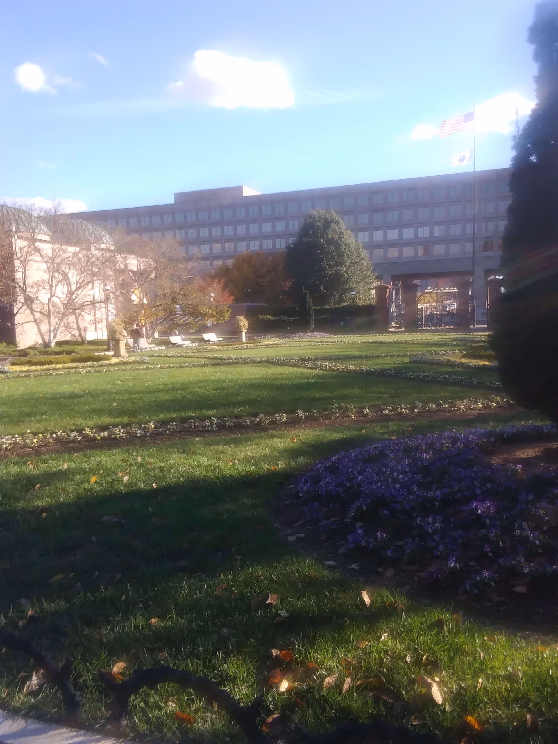 A landscaped outdoor garden area with green grass, flower beds, trees, and a large building in the background under a blue sky with some clouds. There are also two flagpoles with flags flying near the building.