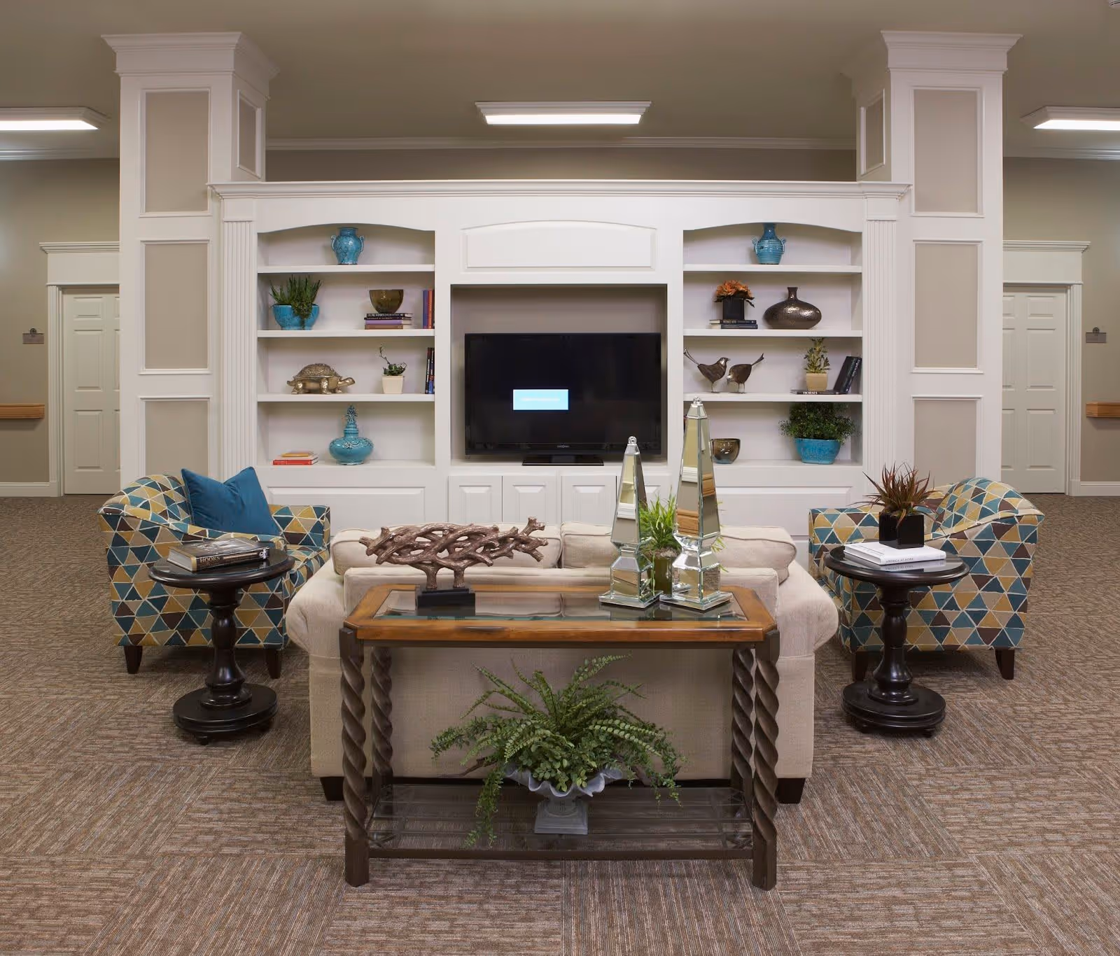 A cozy living room area in a senior living facility featuring a beige sofa with decorative pillows, two patterned armchairs with blue and yellow geometric designs, and two round black side tables with books and decor. Behind the sofa is a wooden console table with plants and decorative items. The background has built-in white shelves with various decorative vases, plants, and books, and a flat-screen TV mounted in the center. The room has beige carpet and neutral-colored walls with white trim.