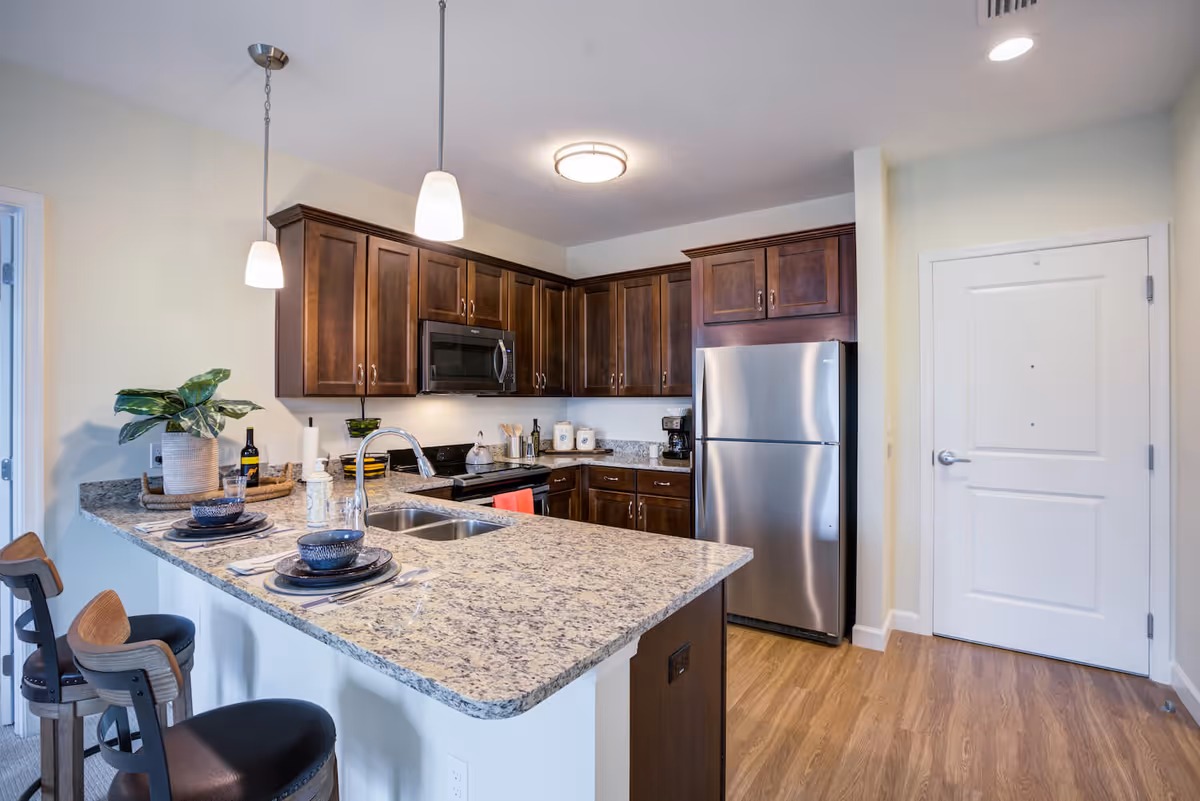 Modern kitchen with granite countertops, dark wooden cabinets, stainless steel refrigerator and microwave, a double sink, and two pendant lights hanging above the counter with two bar stools set with plates and bowls.