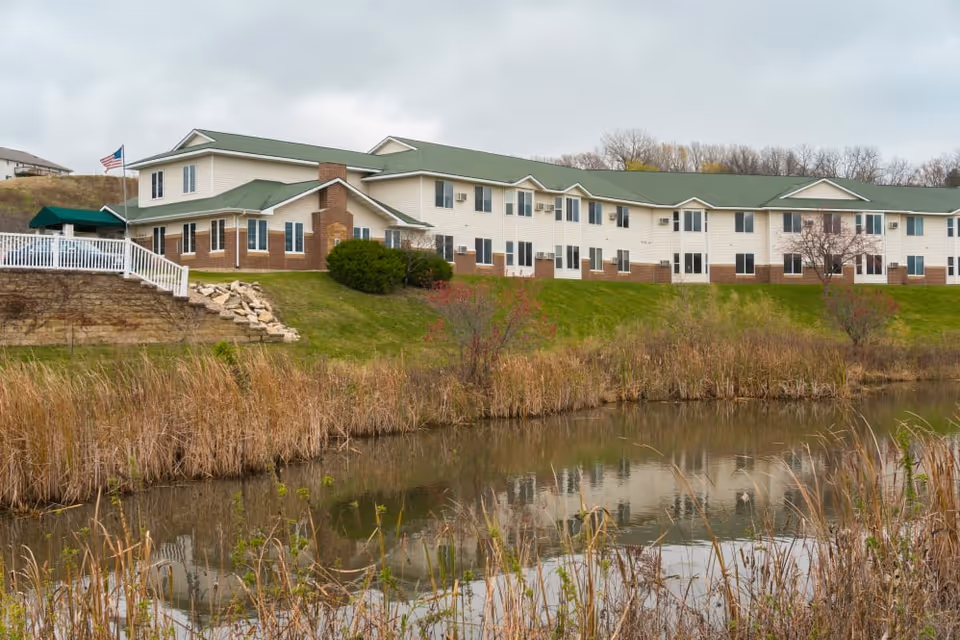 Exterior view of a two-story senior living facility building with a green roof and beige siding, situated on a grassy hill next to a pond with tall reeds and vegetation. An American flag is visible near the building.