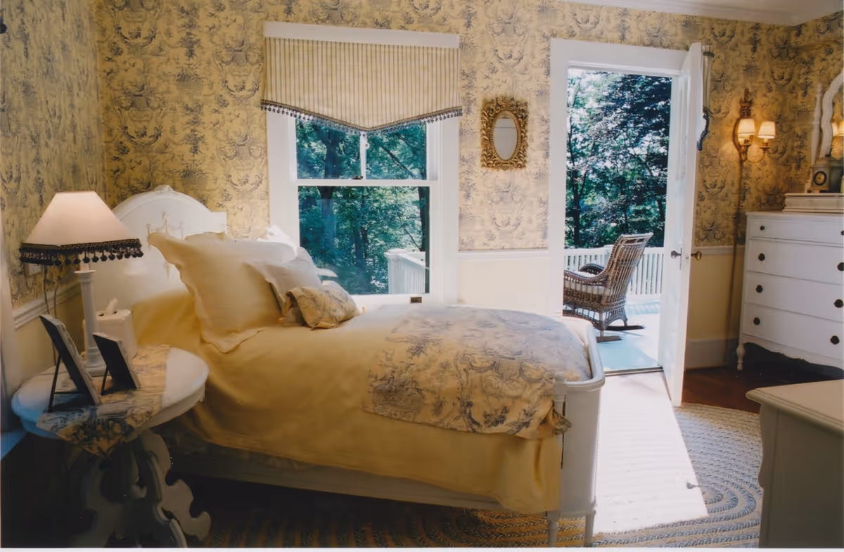 Cozy traditional bedroom with a bed, nightstand and lamp, dresser, and an open door leading to a wooded porch with a wicker chair.