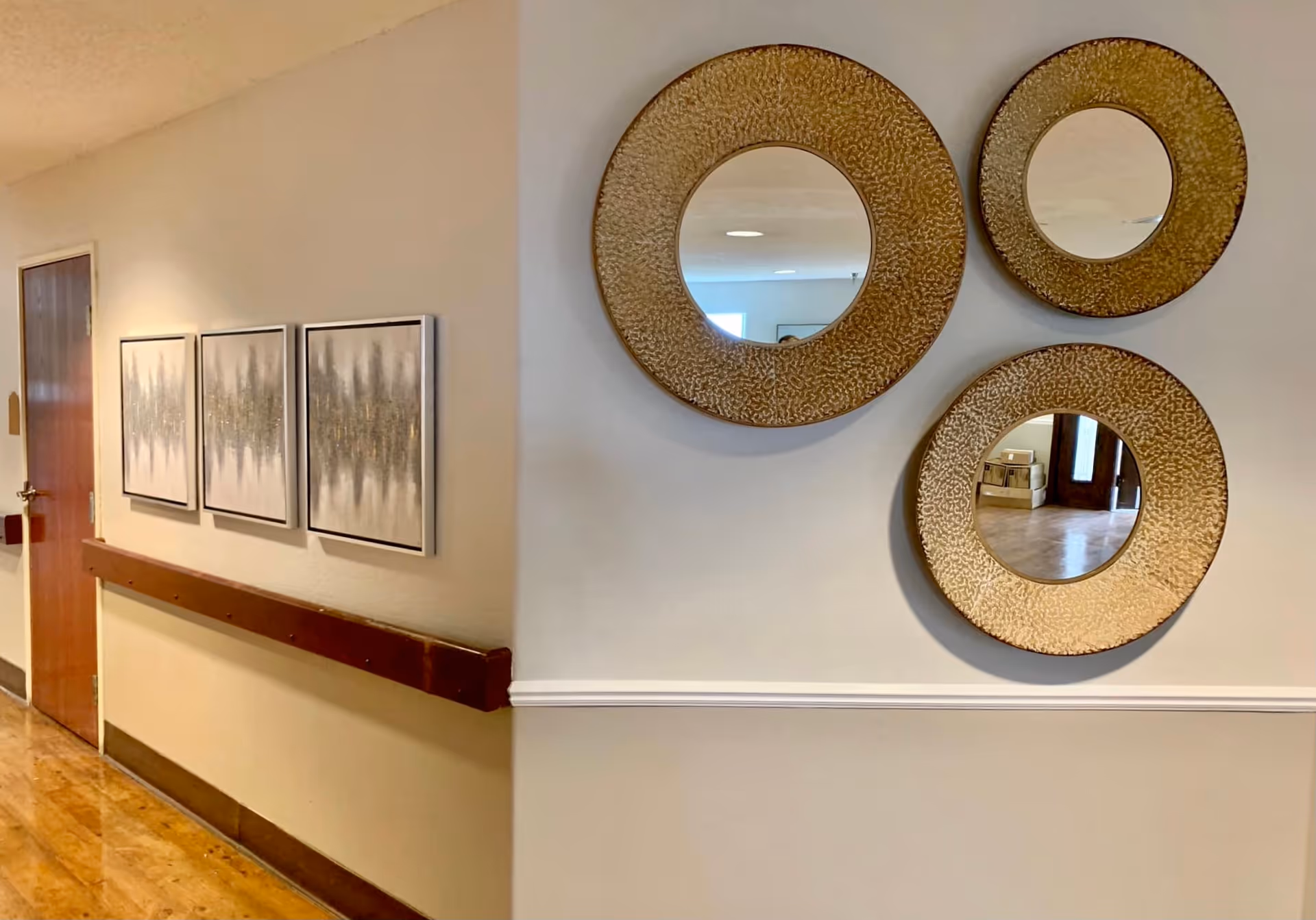 Interior hallway of a senior living facility with a wooden door on the left, three framed abstract art pieces on the left wall, and three decorative round mirrors with textured golden frames on the right wall. The floor is polished wood, and there is a wooden handrail along the left wall.