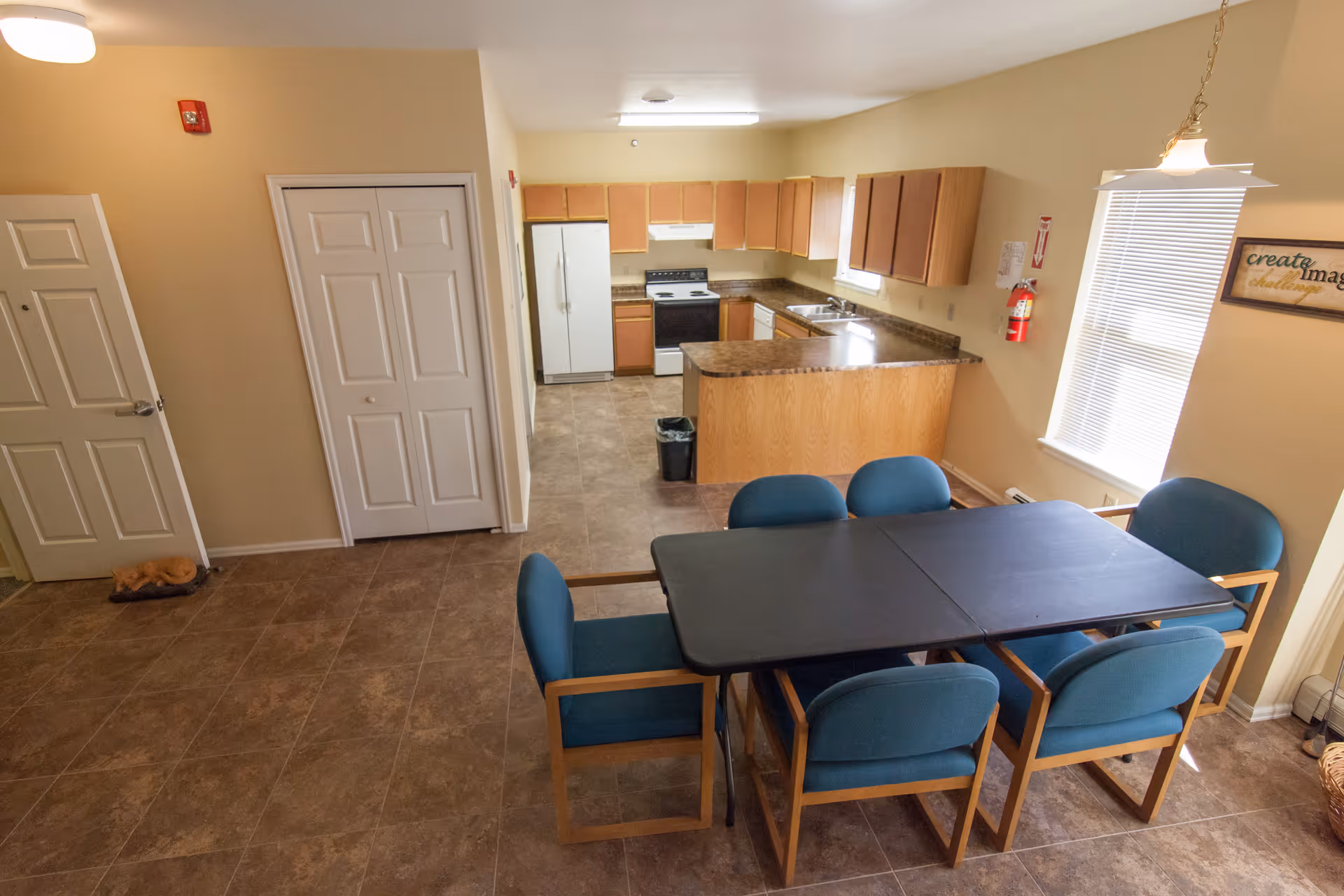 Interior view of a kitchen and dining area in a senior living facility. The kitchen features wooden cabinets, a white refrigerator, stove, and dishwasher. A black dining table with six blue cushioned chairs is in the foreground. The room has beige walls, tiled floors, a window with blinds, and a fire extinguisher mounted on the wall.