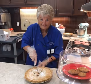 A woman wearing a blue Azura Memory Care of Oak Creek uniform is cutting a pie in a kitchen area. There are baked goods on the counter, including cookies under a glass dome and a tray of cookies in the background. Kitchen appliances and cabinets are visible behind her.