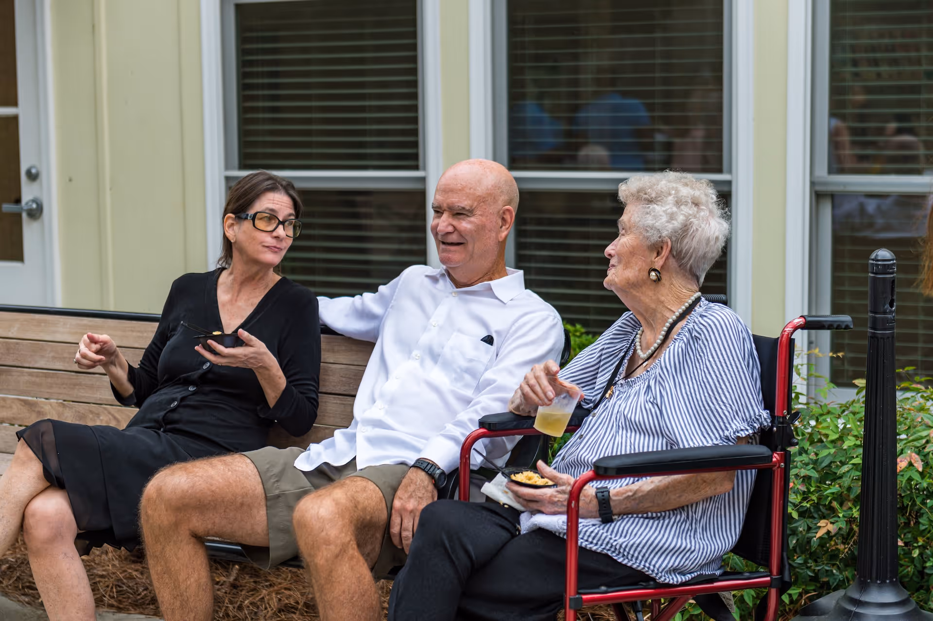 Three people sitting outside on a bench and a wheelchair, engaged in conversation. An elderly woman in a wheelchair holds a drink and a snack, a man in a white shirt and shorts sits next to her, and a woman in a black dress with glasses sits on the bench beside them. They are in front of a building with windows and greenery around.