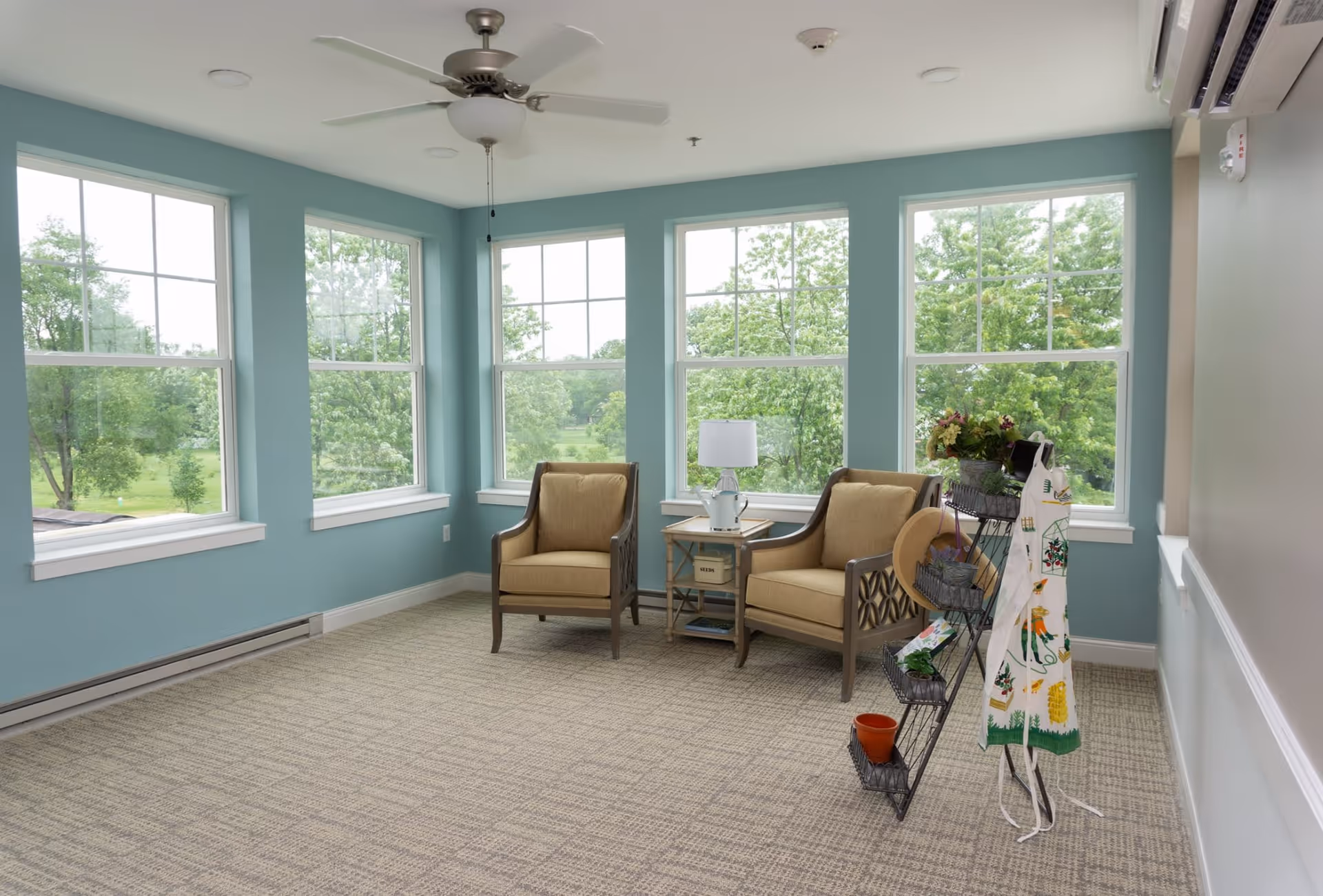A bright corner room with large windows showing green trees outside. The room has light blue walls, beige carpet, two cushioned armchairs with wooden frames, a small side table with a lamp, and a decorative rack holding plants, a hat, and an apron. A ceiling fan is mounted above.