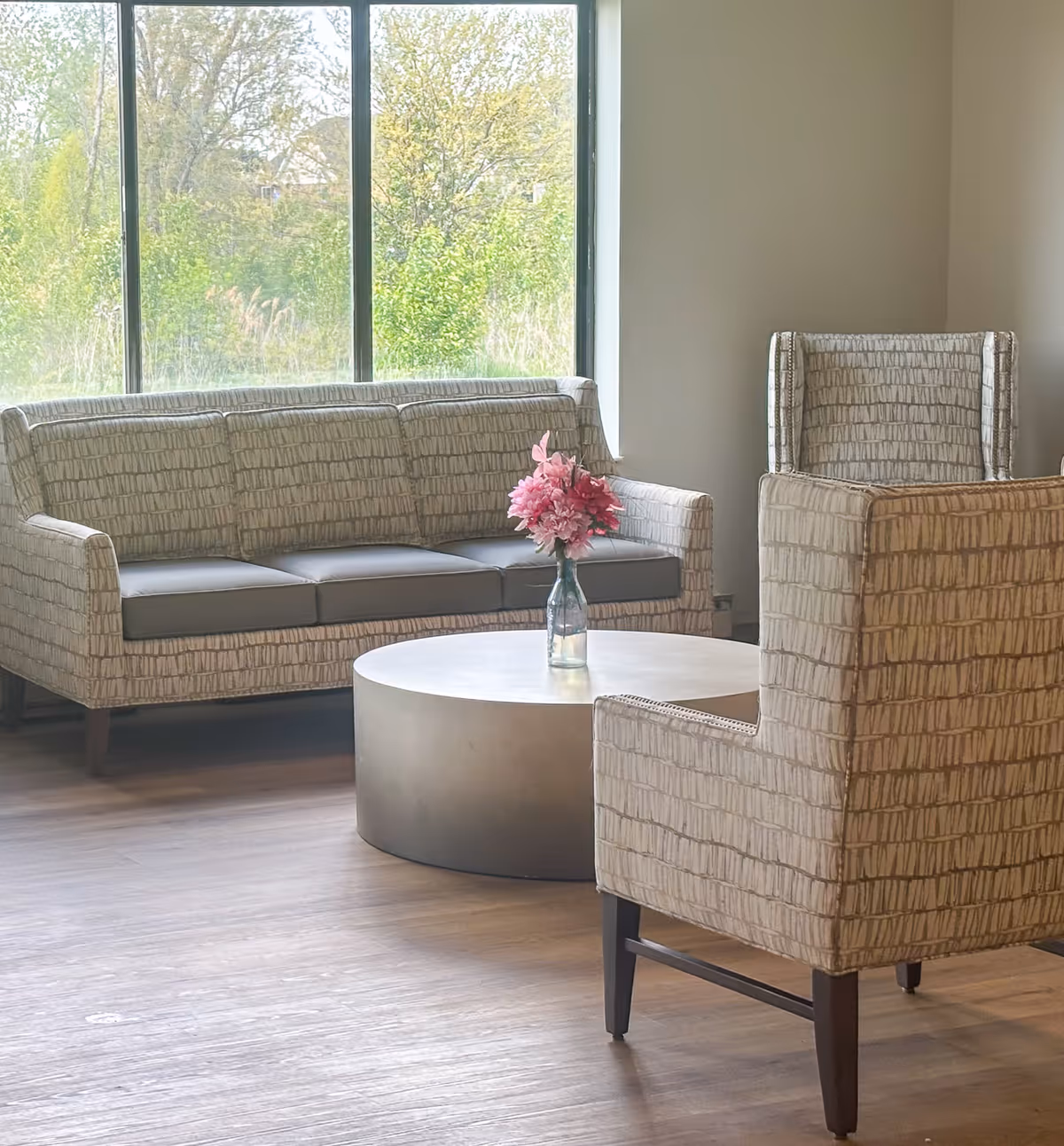 Bright seating area with patterned sofa and armchairs around a round coffee table holding a vase of pink flowers in front of a large window showing greenery.
