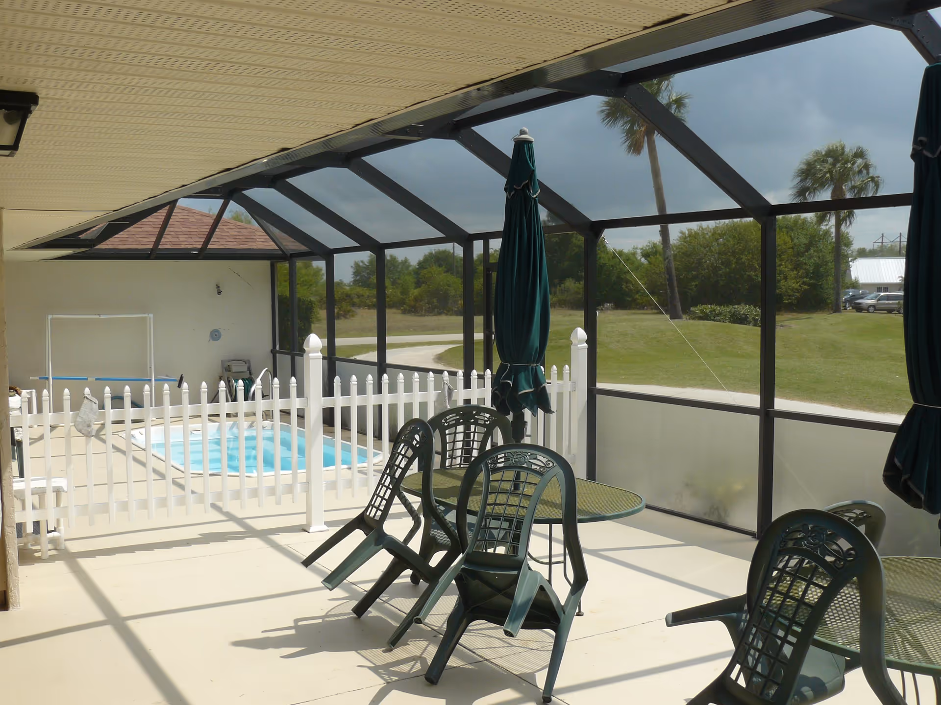 Screened-in patio with plastic chairs and tables, closed umbrellas and a white picket fence around a small swimming pool overlooking a grassy yard.