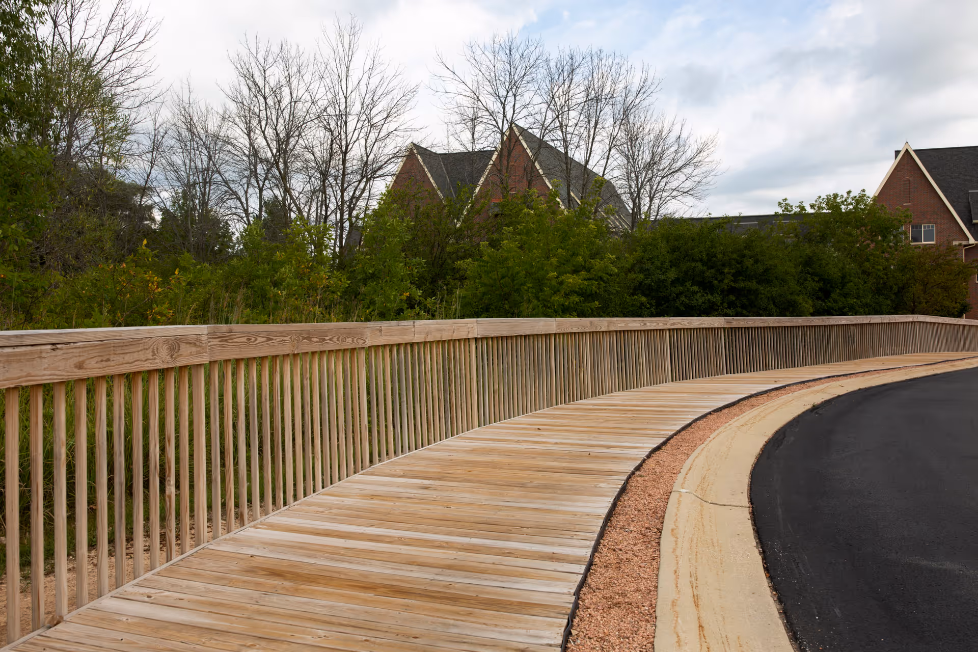 Curved wooden boardwalk with a railing beside a paved road, trees and brick buildings visible in the background.