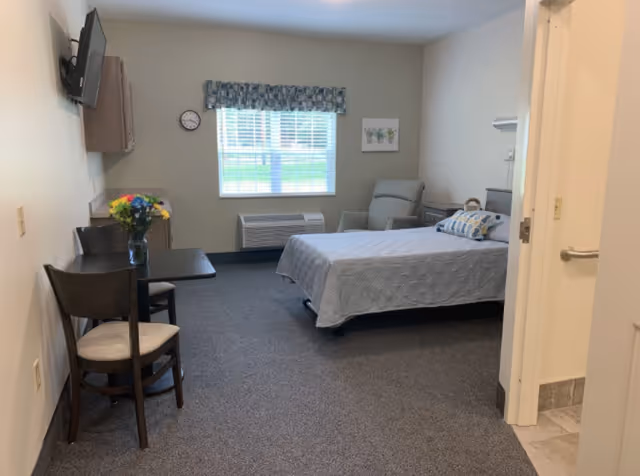 A senior living facility bedroom with a bed covered in a gray quilt, a cushioned armchair, a small table with two chairs, a wall-mounted TV, a window with blinds and a floral valance, a clock on the wall, and a small piece of artwork. The room has carpeted flooring and neutral-colored walls. A bathroom entrance is visible on the right side.