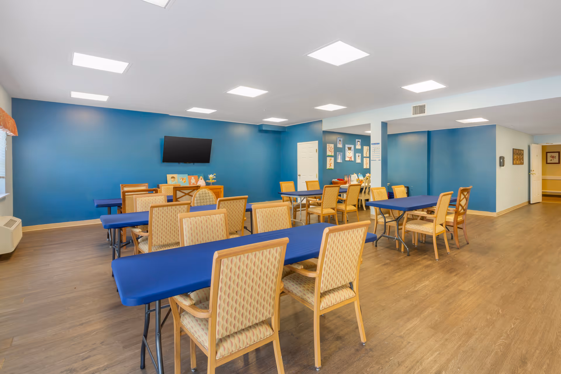 A spacious room with several tables covered in blue tablecloths and multiple chairs arranged around them. The walls are painted blue and white, with a flat-screen TV mounted on one blue wall. The floor is wooden, and the ceiling has multiple square recessed lights. There are framed pictures and decorations on the walls, and a door is visible in the background.
