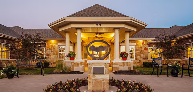 Exterior view of Silverado Overland Park Memory Care Community building at dusk, featuring a stone facade, a covered entrance with columns, a circular metal sculpture in front, and landscaped flower beds and benches on either side.