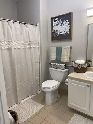 A clean bathroom with a white toilet, a shower with a beige curtain, a white sink with a countertop, and a basket with towels. Above the toilet, there is a towel rack with two folded towels and a framed floral artwork on the wall.