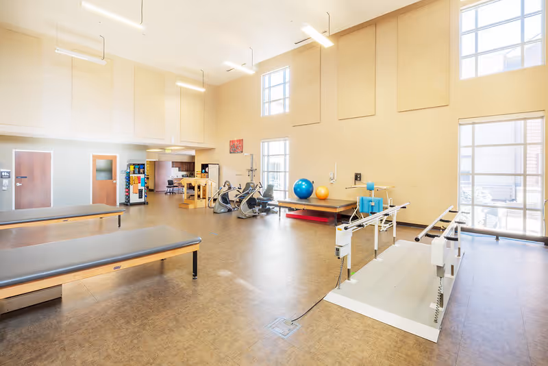 A spacious physical therapy or rehabilitation room with high ceilings and large windows allowing natural light. The room contains therapy tables, exercise bikes, therapy balls, and parallel bars for walking exercises. The walls are light-colored, and there is a vending machine and a restroom door visible in the background.