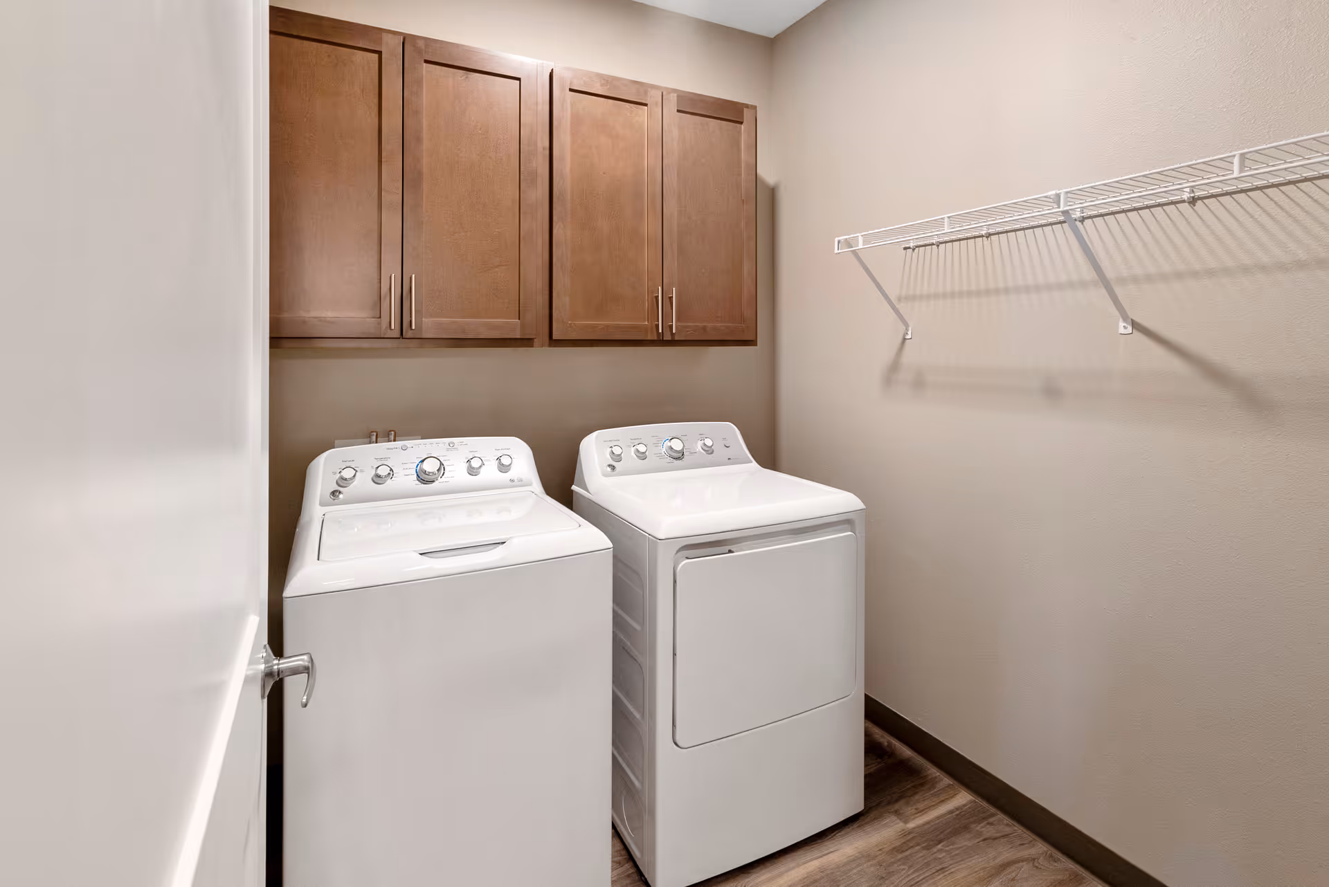 Small laundry room with a white top-loading washer and matching dryer beneath wood cabinets and a wire shelf.
