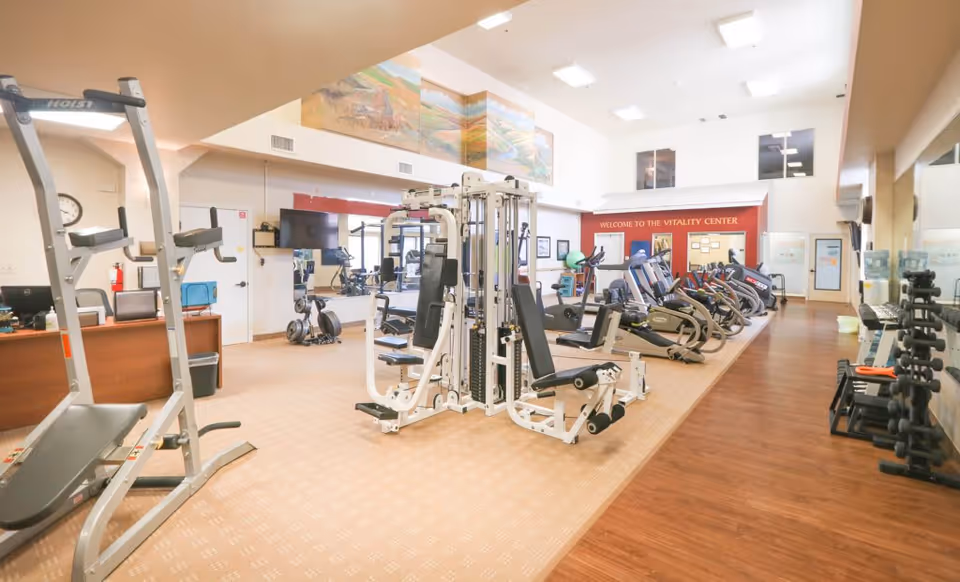 A spacious fitness center with various exercise equipment including weight machines, treadmills, stationary bikes, and free weights. The room has high ceilings, large windows, and a sign that reads 'Welcome to the Vitality Center'. The floor is a combination of carpet and wood, and there is a mural on the upper wall.