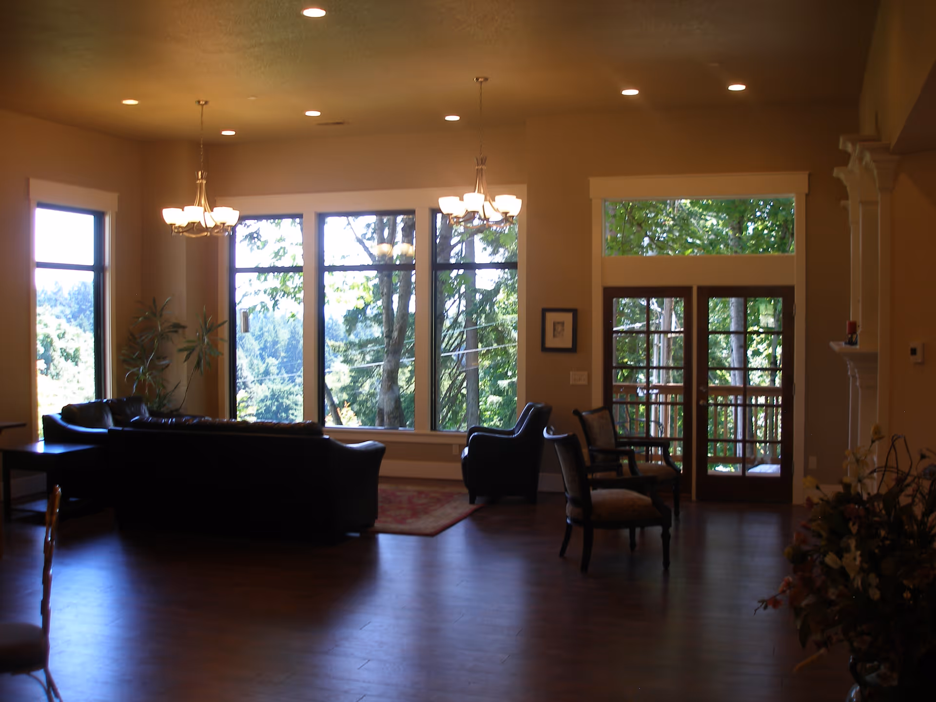 A spacious living room with large windows showing a view of trees outside. The room has dark leather sofas, two upholstered chairs, a red area rug, wooden flooring, and two chandeliers hanging from the ceiling. There is a glass door leading to an outdoor area and a fireplace on the right side with a floral arrangement nearby.