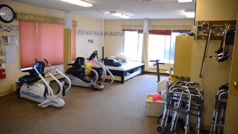 A bright therapy room with exercise machines, treatment beds, and stacked walkers and mobility aids.