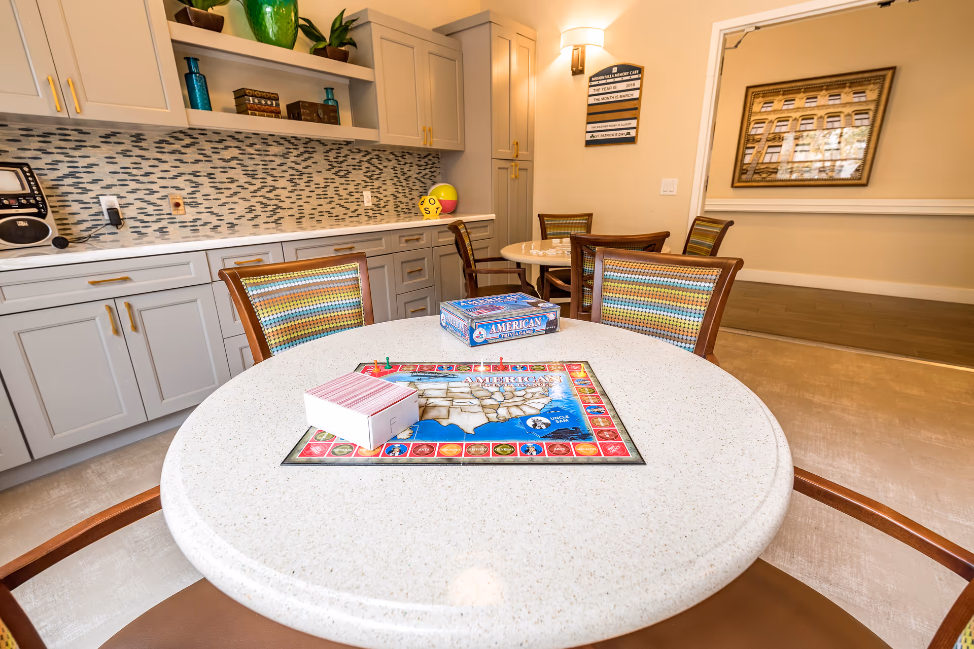 A bright activity room with a round table in the foreground featuring an American trivia board game and cards. The room has light-colored cabinetry with gold handles, a mosaic tile backsplash, and several chairs with multicolored striped upholstery. In the background, there is another round table with chairs and a wall-mounted board displaying the date and weather information.