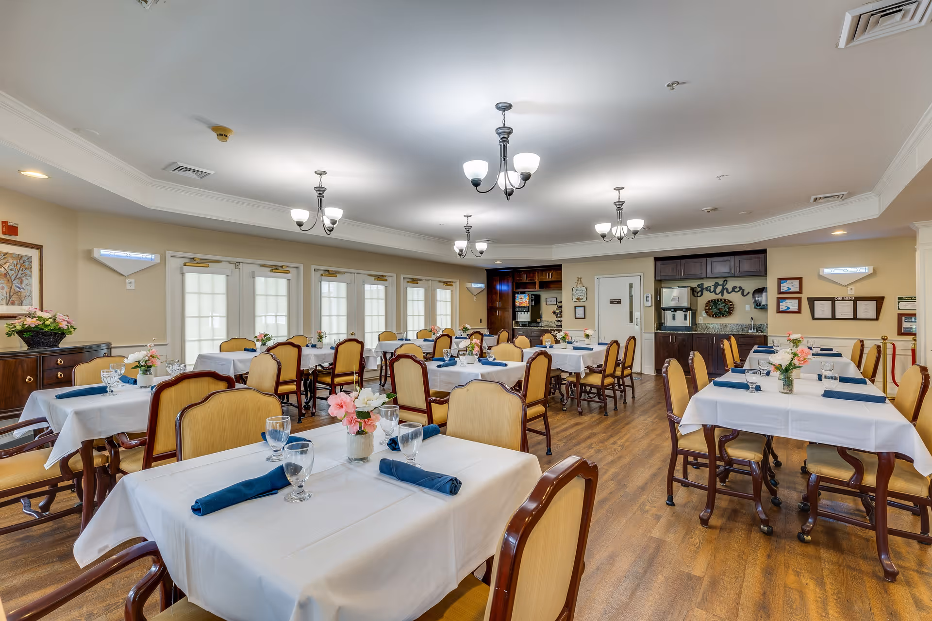 A bright and spacious dining room with multiple tables covered in white tablecloths, each set with glassware, blue napkins, and small floral centerpieces. The room features wooden chairs with cushioned seats, hardwood flooring, and several ceiling light fixtures. Large windows with white curtains line one wall, and a small kitchenette area is visible in the background with a sign that says 'Gather'.
