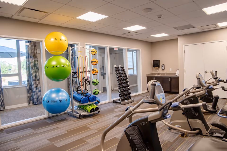 A well-lit fitness room with exercise equipment including stationary bikes, a rack of dumbbells, colorful exercise balls, resistance bands, and yoga mats. Large windows with curtains allow natural light to enter, and there is a small sink and cabinet area in the corner.