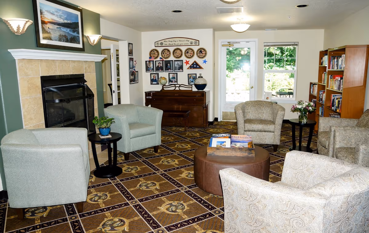 Cozy senior living lounge with armchairs arranged around a central ottoman, a fireplace, piano, bookshelves and a door to an outdoor patio.