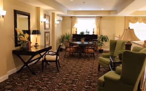 A cozy living room area with patterned carpet, green upholstered armchairs, a wooden table with chairs, a console table with a mirror and lamp, and several plants near windows with curtains.