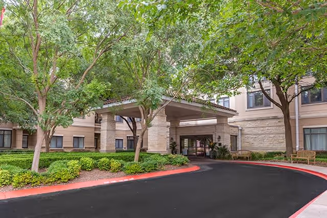 Entrance to a senior living facility with a covered drop-off area, surrounded by trees and landscaped bushes, with benches near the building's windows.
