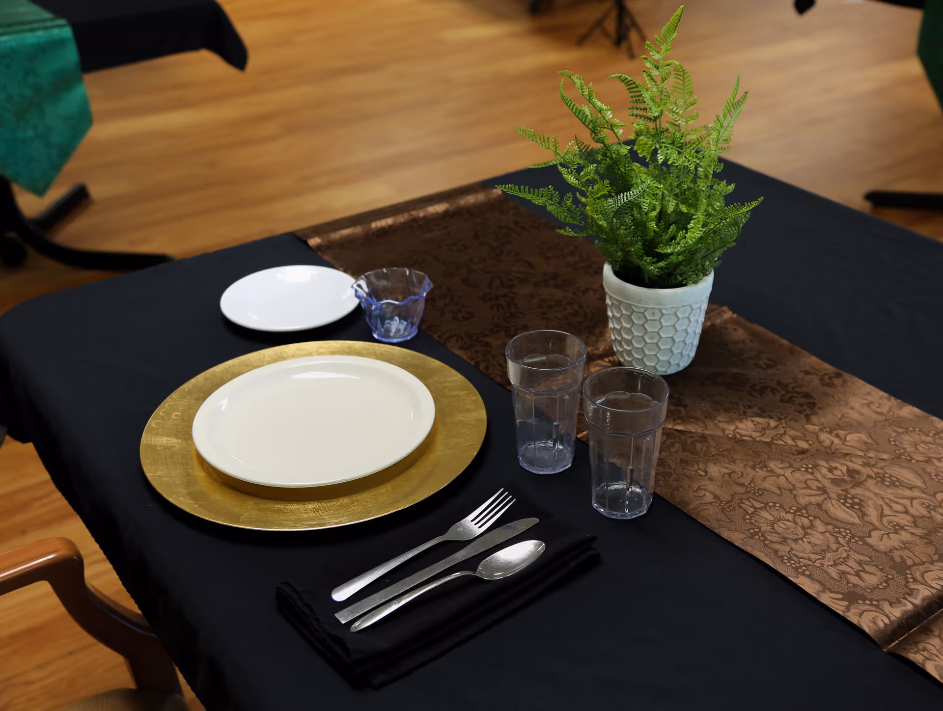 A table set with plates, cutlery, two glasses and a potted fern on a black tablecloth with a brown runner in a dining area.