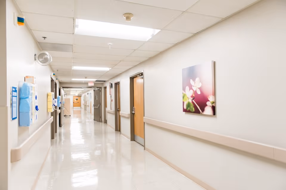 A clean, well-lit hospital hallway with beige walls and a shiny tiled floor. Several closed wooden doors line the corridor on the right side, and handrails run along both walls. A framed picture of white flowers on a pink background hangs on the right wall. Medical supplies and informational materials are mounted on the left wall.