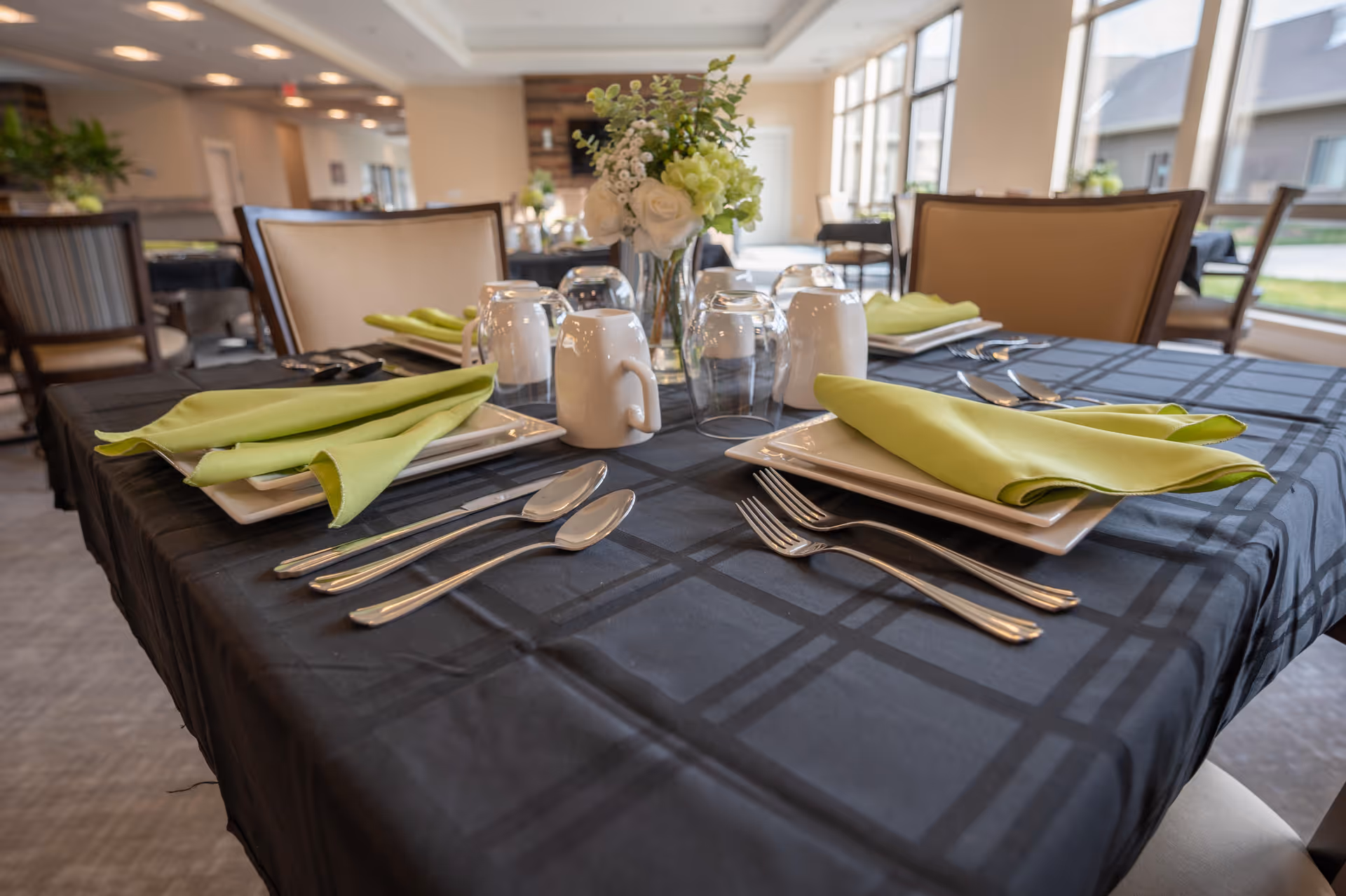 A dining table set for four with black checkered tablecloth, white square plates, green folded napkins, silverware, white mugs, upside-down glasses, and a floral centerpiece in a bright dining room with large windows and beige chairs.