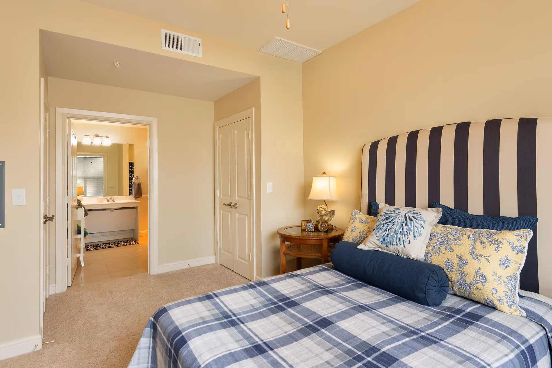 A bedroom with a bed featuring a blue and white plaid bedspread and multiple decorative pillows. The headboard is striped in dark blue and beige. Next to the bed is a round wooden nightstand with a lamp and small decorative items. The room has beige walls and carpeted floor. An open door reveals a bathroom with a sink and mirror.