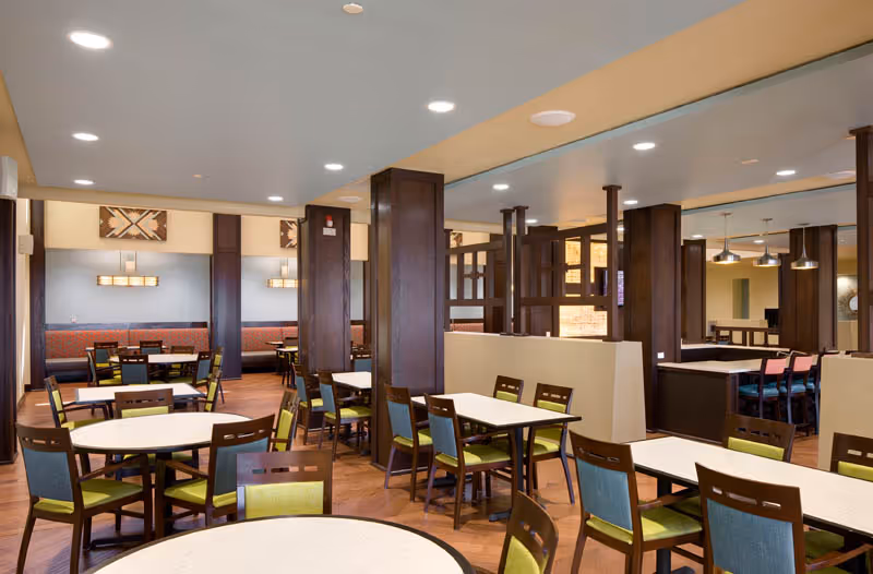 Spacious communal dining room with round and rectangular tables and wooden chairs with green and blue cushions, warm wood pillars, and pendant lighting.