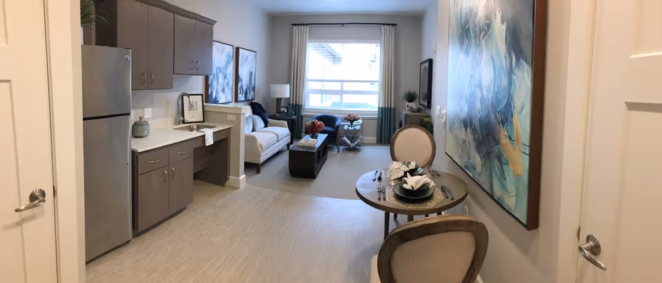 A bright and modern living space featuring a small kitchen area with gray cabinets and a stainless steel refrigerator on the left, a cozy living room with a white sofa, two blue chairs, a coffee table, and a large window with curtains in the background. On the right, there is a round dining table set for two with elegant place settings and a large abstract painting on the wall.