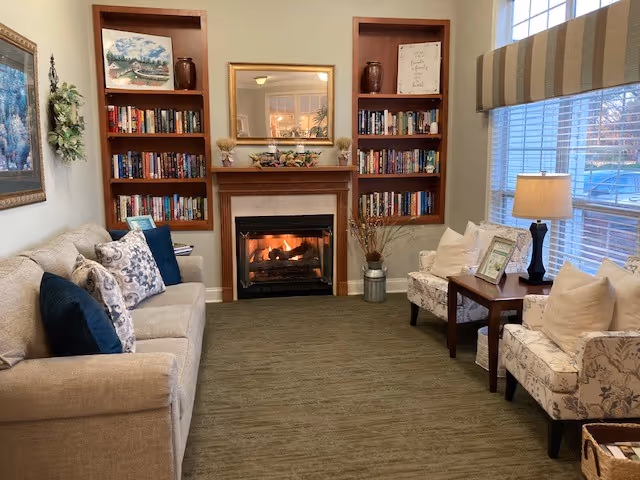 A cozy living room with a beige sofa on the left adorned with patterned and solid pillows, two floral armchairs on the right with a wooden side table and lamp between them, a fireplace with a fire burning in the center flanked by built-in bookshelves filled with books and decorative items, a large window with striped valance on the right, and a mirror above the fireplace reflecting a chandelier.